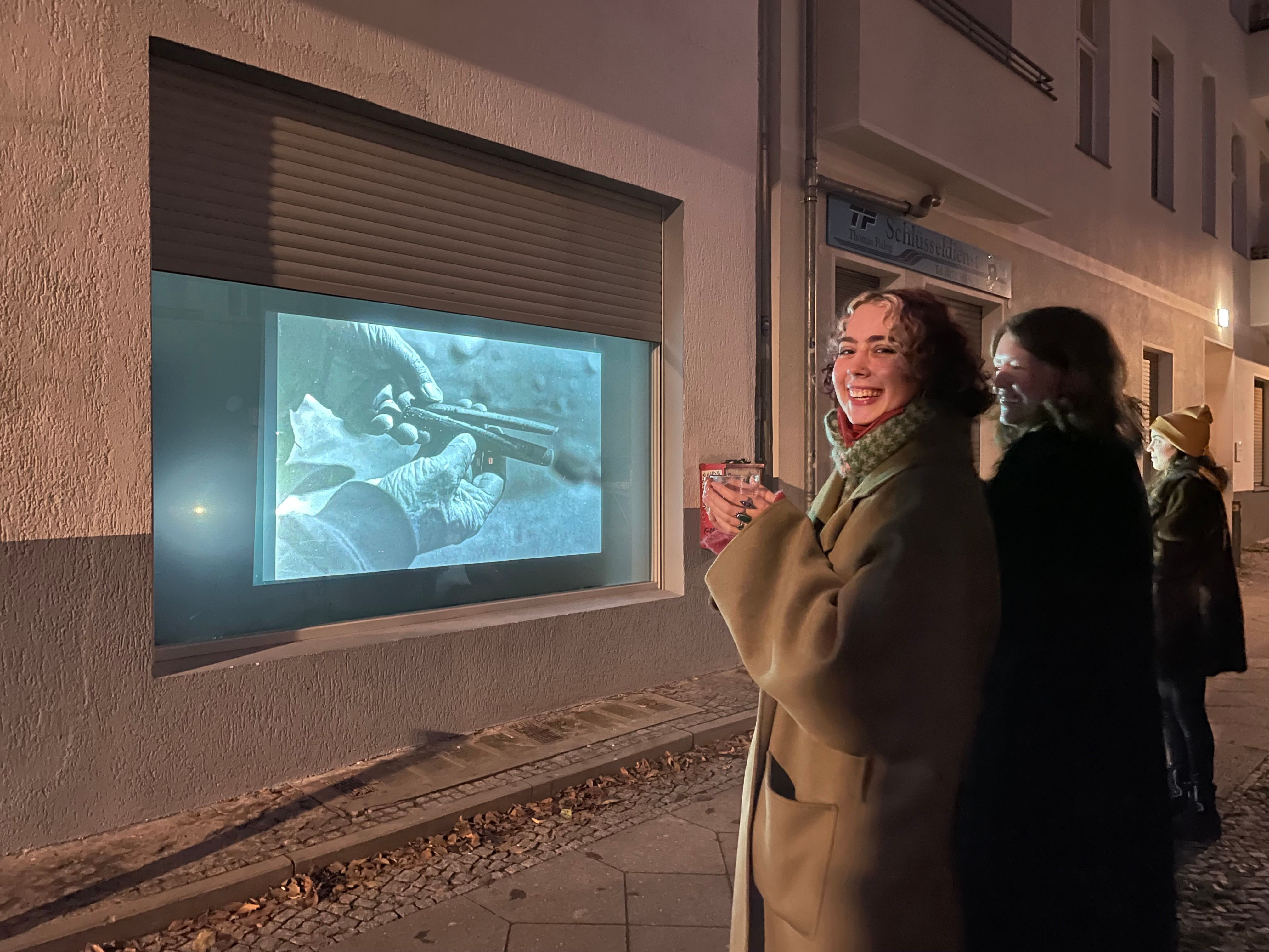 Two students smile at the camera, on the sidewalk in front of a projected photograph of gloved hands.