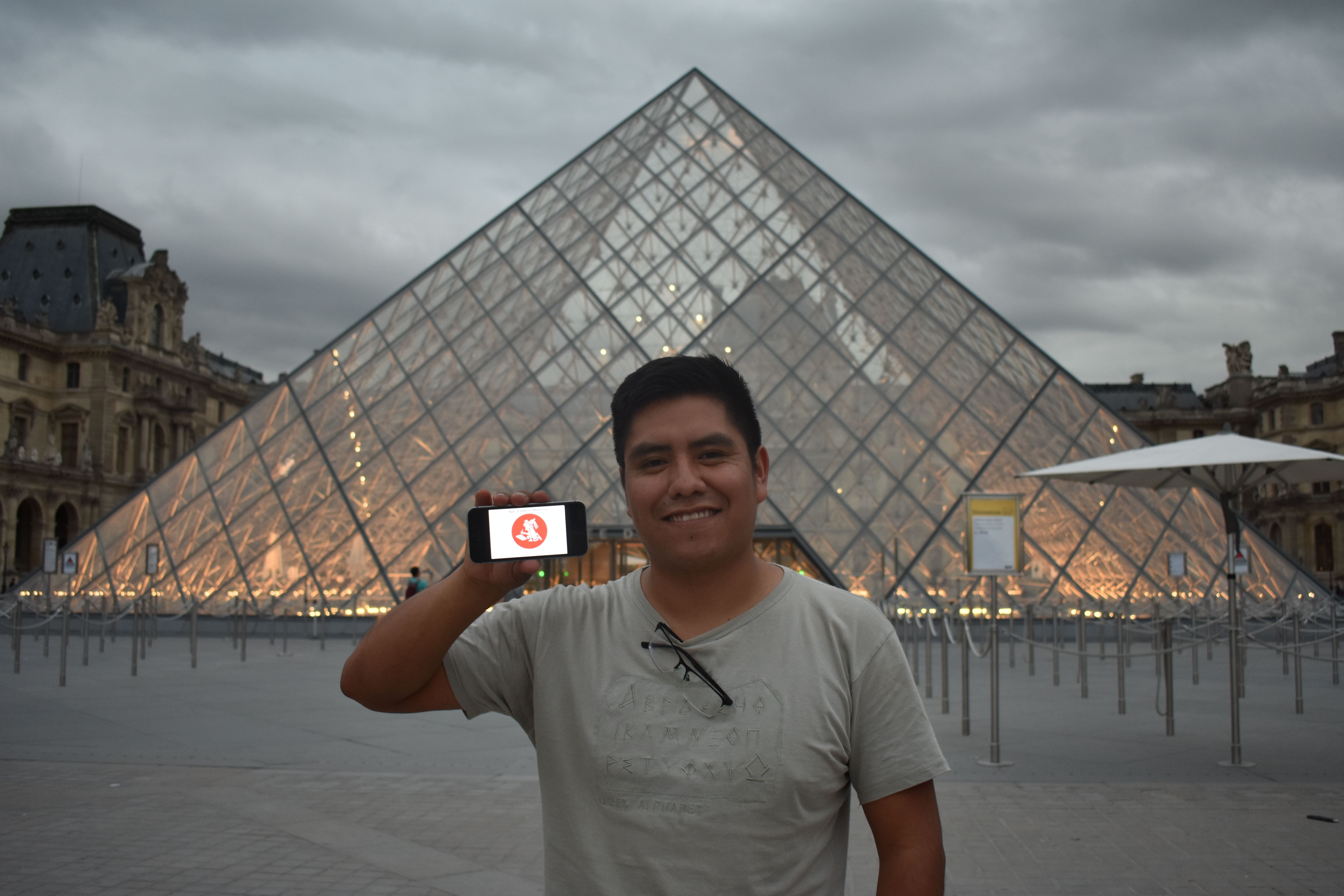 Aguilar poses in front of a glass pyramid building, holding up his phone with a graphic