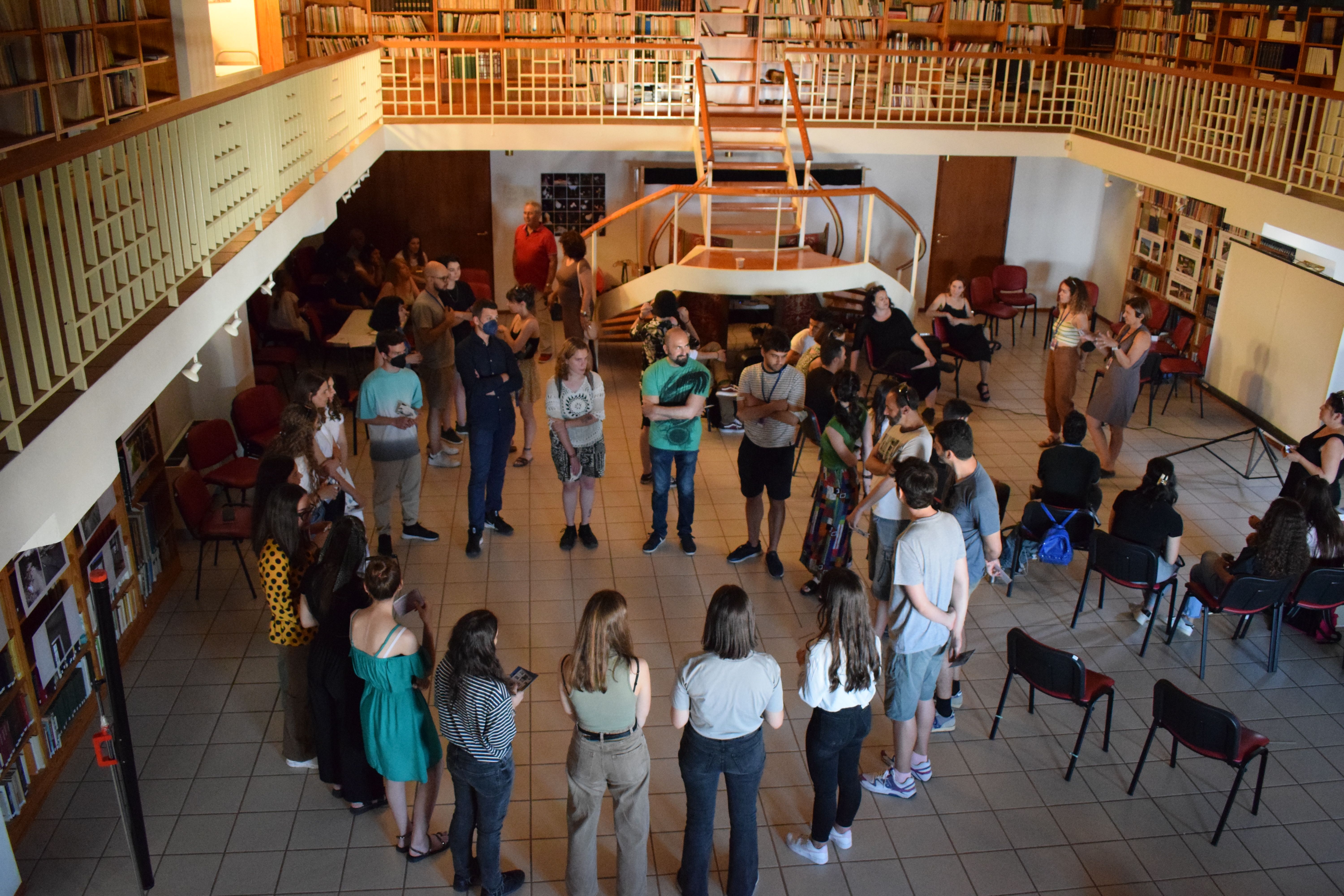 Group of people standing in a circle in a large room with an upstairs library balcony.