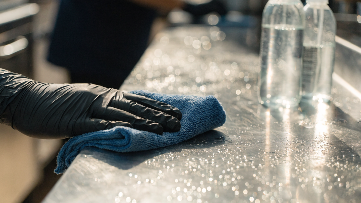 A gloved hand wiping a stainless-steel counter with a blue microfiber cloth in a commercial kitchen, spray bottles nearby.