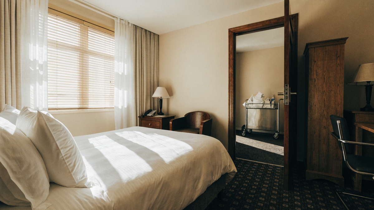 A pristine sunlit hotel guest room with crisp white linens and a housekeeping cart in the open doorway.