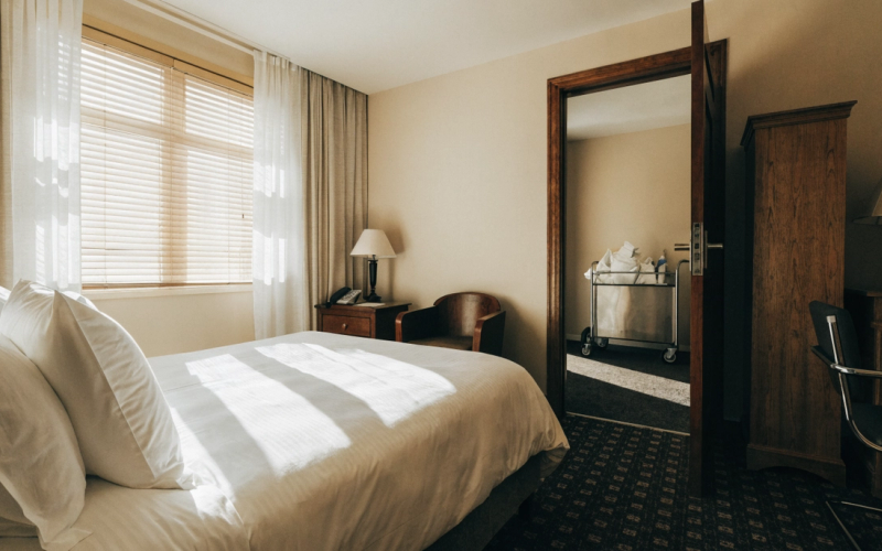 A pristine sunlit hotel guest room with crisp white linens and a housekeeping cart in the open doorway.