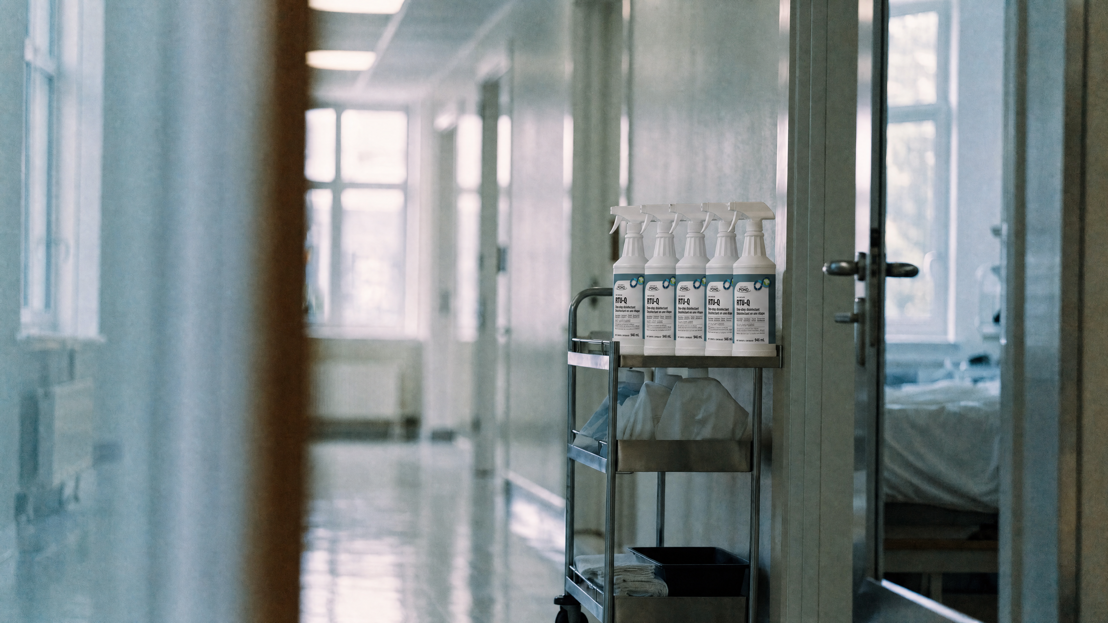 A cleaning cart with neatly arranged spray bottles parked in a bright, freshly cleaned hospital corridor.