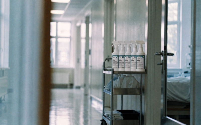 A cleaning cart with neatly arranged spray bottles parked in a bright, freshly cleaned hospital corridor.