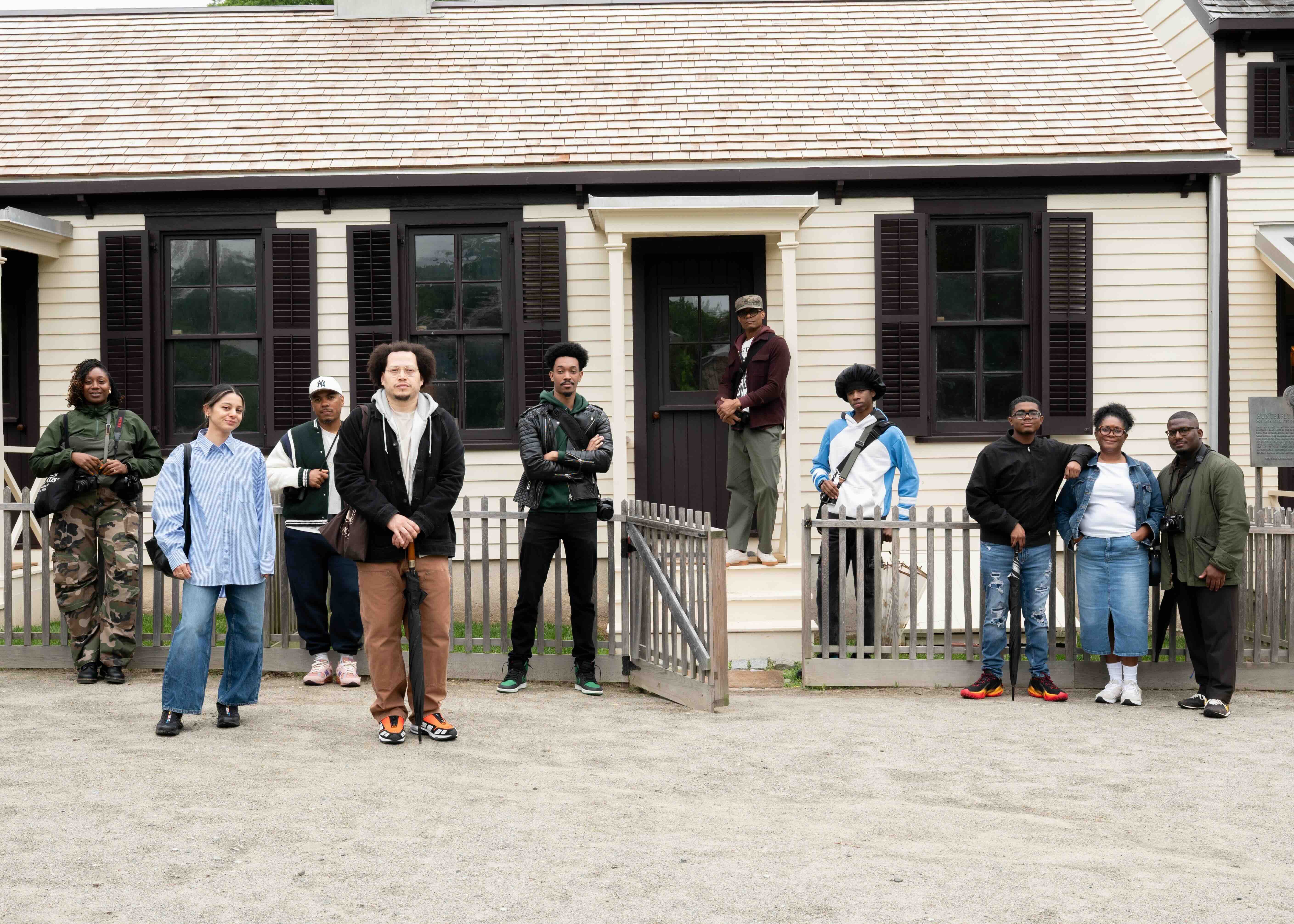 A group of 10 people, a mix of men and women, are standing in front of a light-colored, clapboard building with a shingled roof. The building has dark shutters on its windows and a dark-colored door. A light-colored wooden fence runs horizontally across the foreground, with an open gate in the center. The ground is covered in light-colored gravel.