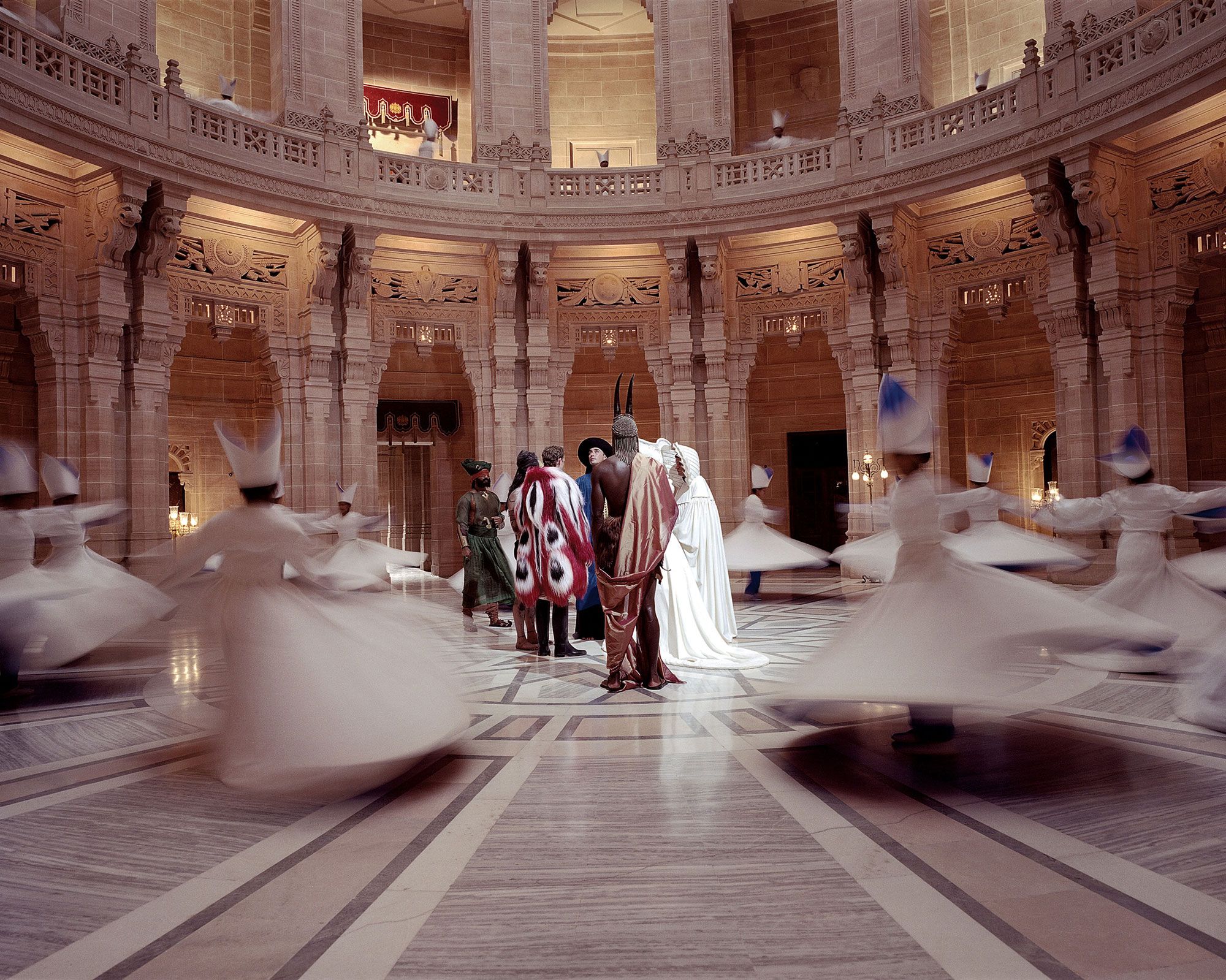 The Fall. 2006. People in elaborate costumes stand in a grand, circular hall while blurred figures in white robes spin around them.