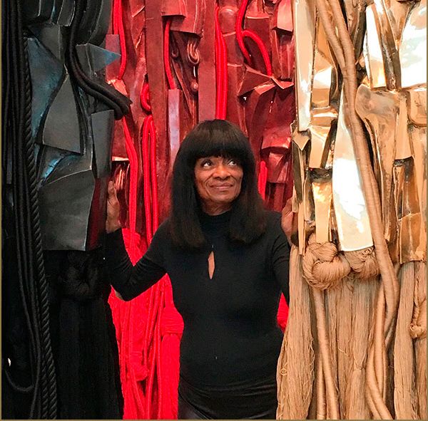 A Black woman with a fringe haircut smiles as she poses between abstract sculptures. The sculptures feature bold textures and colors like black, red, and gold.