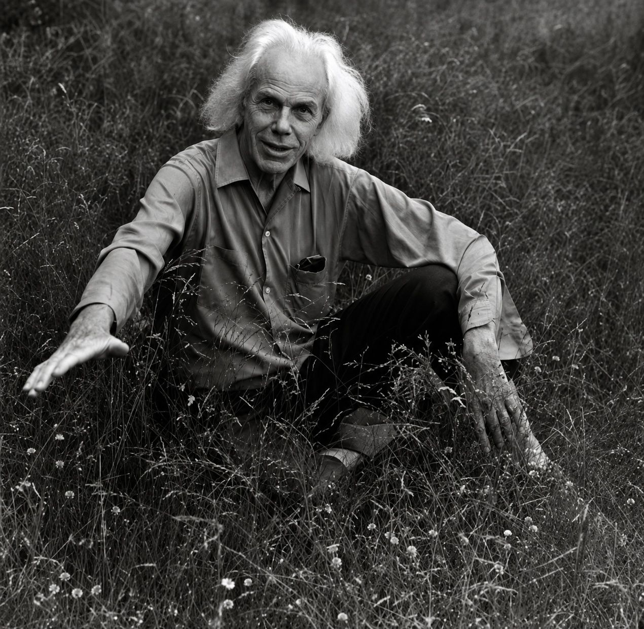 A black and white medium shot shows an elderly man with wild white hair sitting in tall grass. He wears a collared shirt and dark pants, with his left hand outstretched towards the viewer and his right hand resting on his knee.