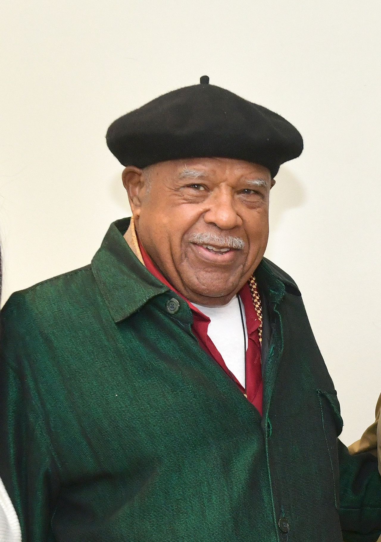 A smiling older Black man wearing a black beret and a green jacket over a red shirt. He is looking directly at the camera.