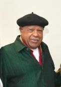 A smiling older Black man wearing a black beret and a green jacket over a red shirt. He is looking directly at the camera.