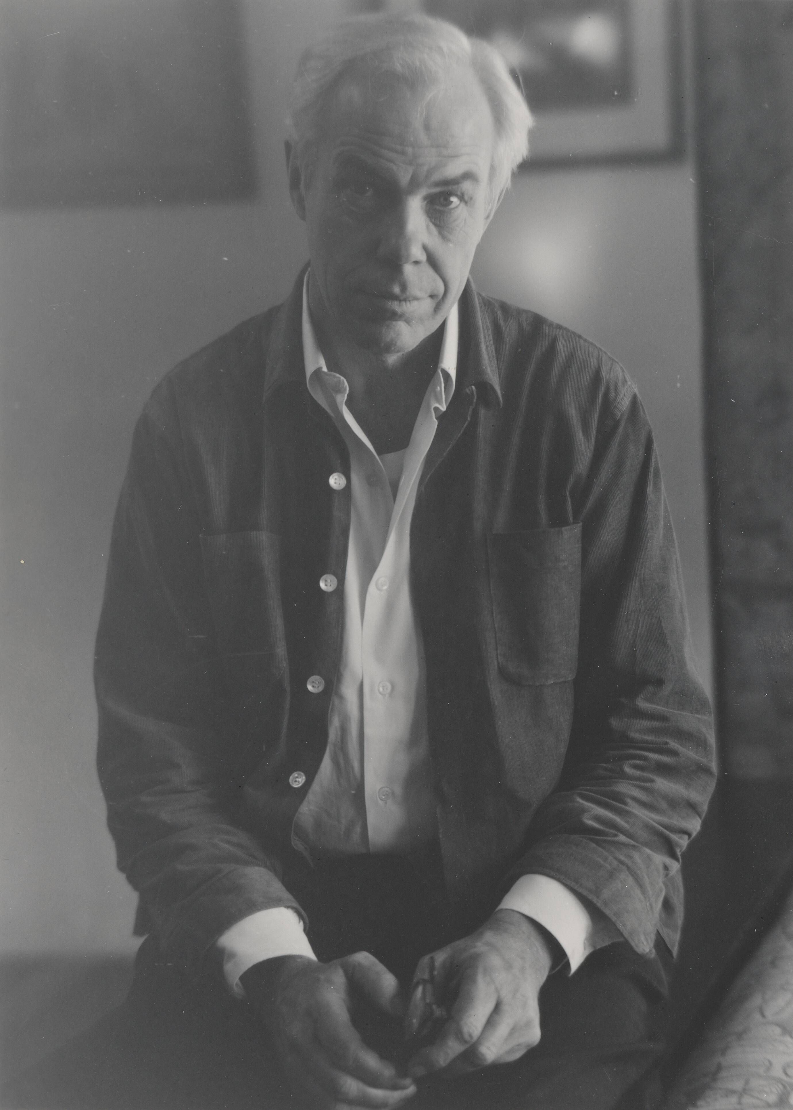 A black-and-white medium shot shows an elderly man with wild white hair sitting in tall grass. He wears a collared shirt and dark pants, with his left hand outstretched toward the viewer and his right hand resting on his knee.