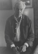 A black-and-white medium shot shows an elderly man with wild white hair sitting in tall grass. He wears a collared shirt and dark pants, with his left hand outstretched toward the viewer and his right hand resting on his knee.