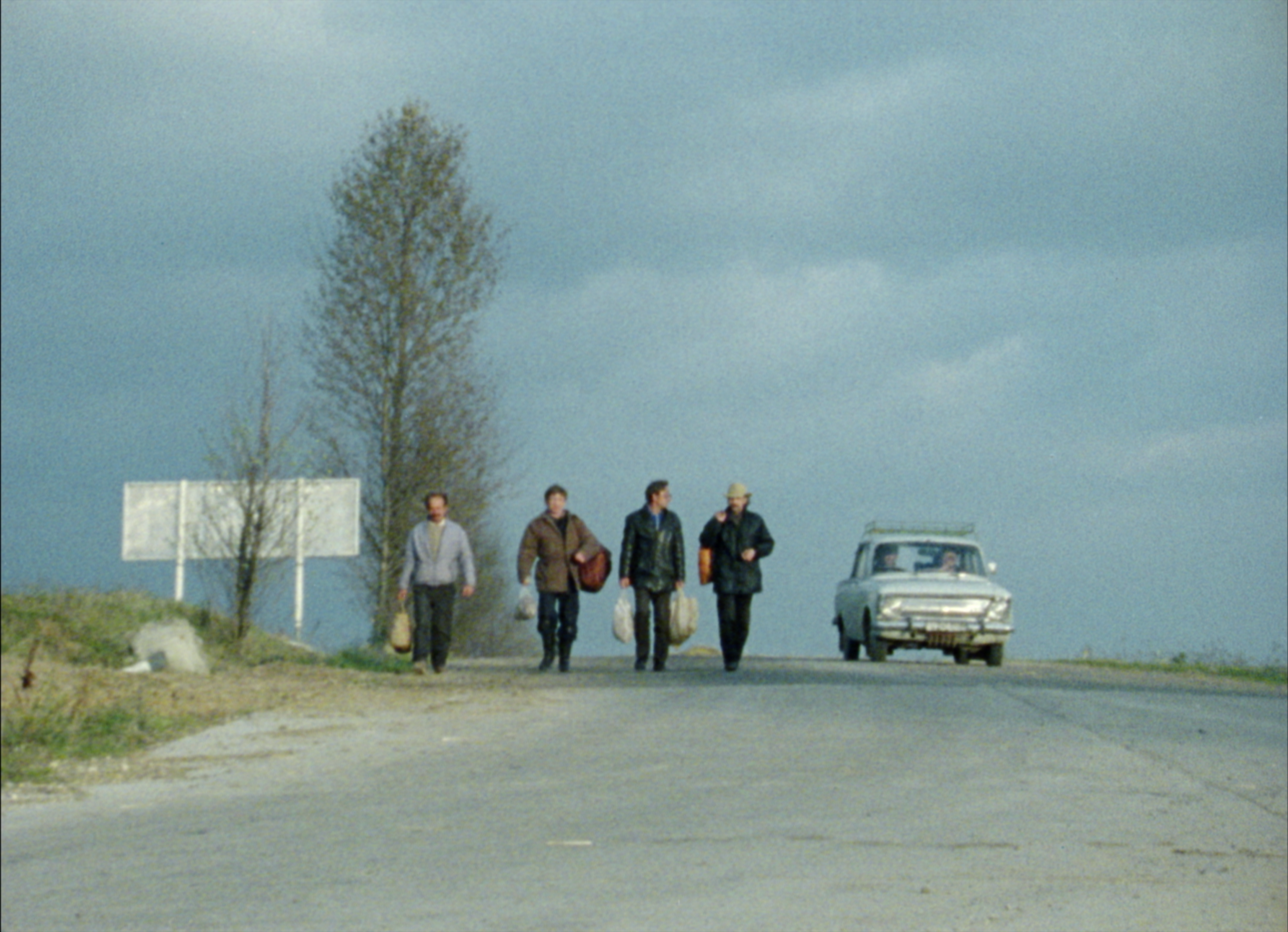 4 men walk down an empty road followed by a car under a gray sky