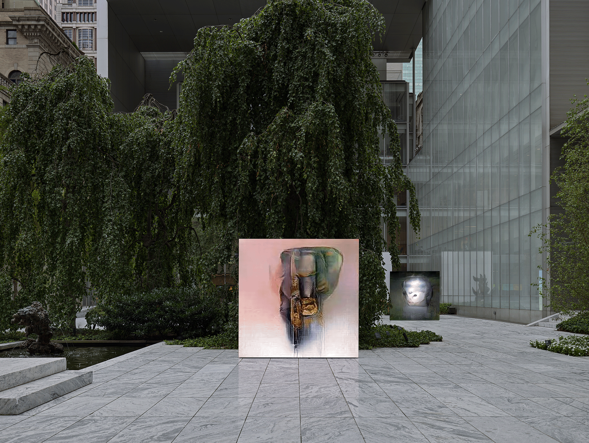 Outdoor museum courtyard with a large weeping tree and ivy-covered walls in the background, glass-walled modern building to the right. In the foreground on a gray marble-paved terrace stands a square freestanding screen or panel displaying a glitchy, pixelated digital image in pink and green tones suggesting an abstract figure or object. A second, smaller dark panel with a glowing white/silver abstract image stands further back to the right, partially obscured by greenery.