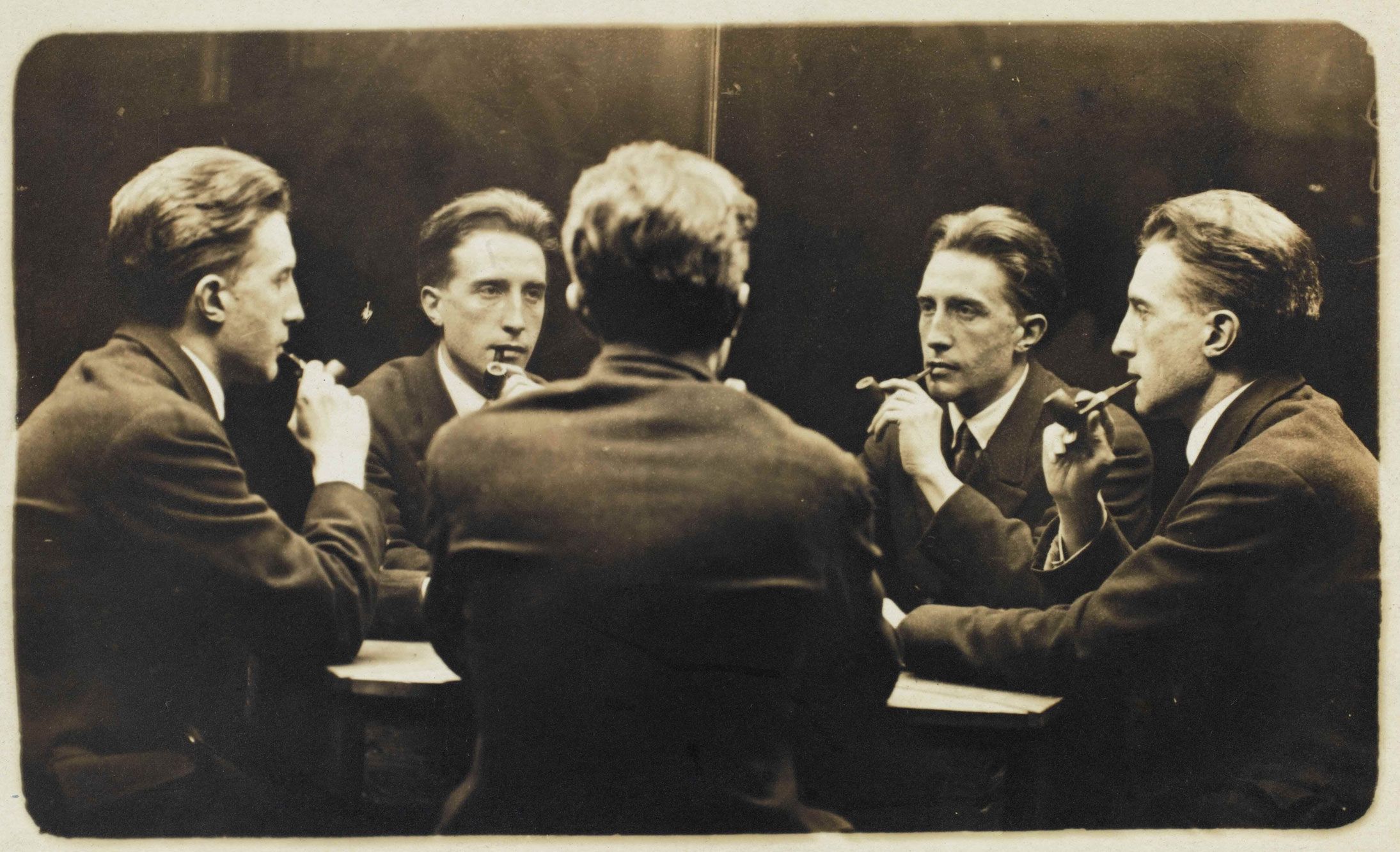 A man with slicked-back hair, smoking a pipe, sits at a table with four different versions of himself. 