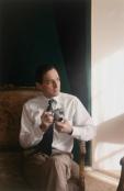 A person in a white shirt and striped tie sits in a chair holding a camera. Predominant colors are white, beige, and dark green.