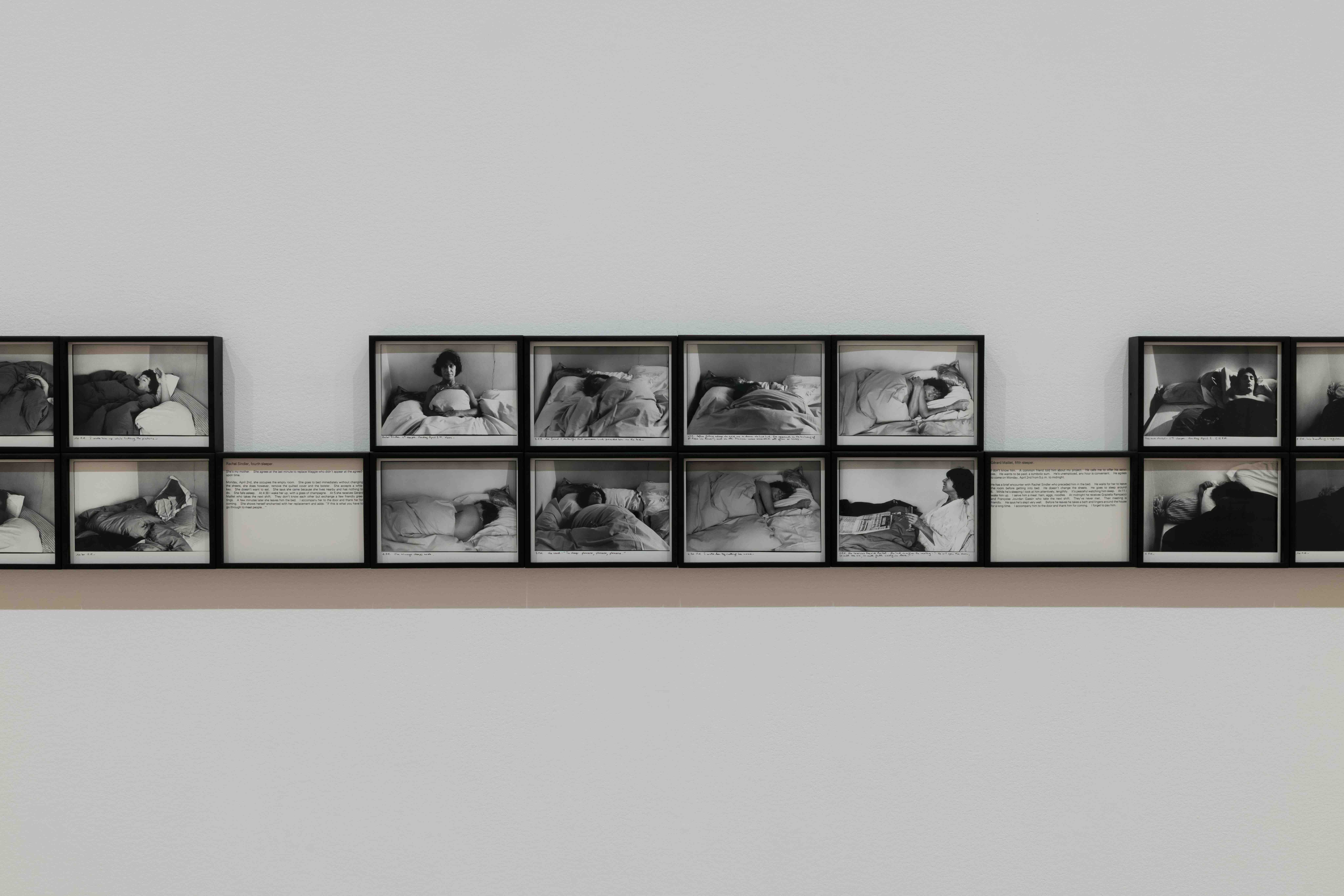 Large installation view showing a grid of many small black-and-white photographs arranged in rows on a wall, each depicting people sleeping or resting in bed