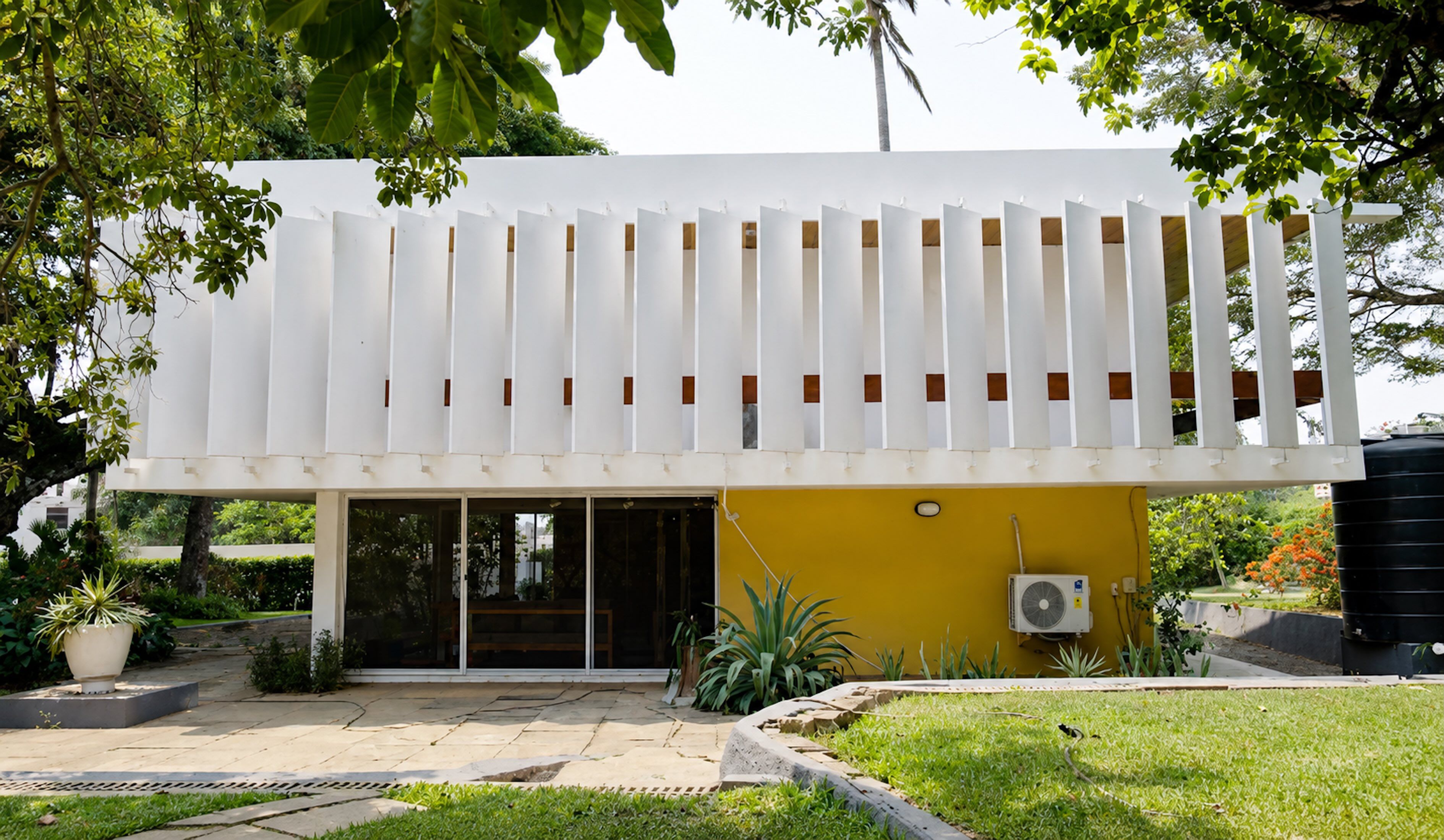 A modern two-story house features a white slatted facade above a yellow lower level with large windows. Lush greenery surrounds the building, with trees and a garden visible.