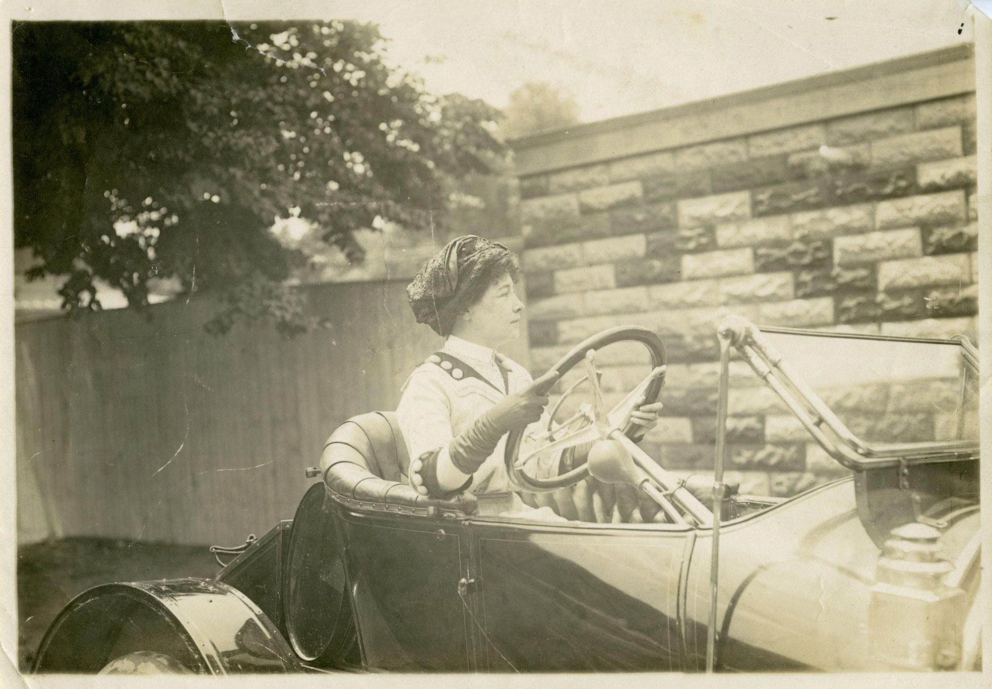 Black-and-white photograph of a woman seated behind the wheel of an early open-top automobile, wearing a hat, gloves, and period clothing, with a stone wall and trees in the background.