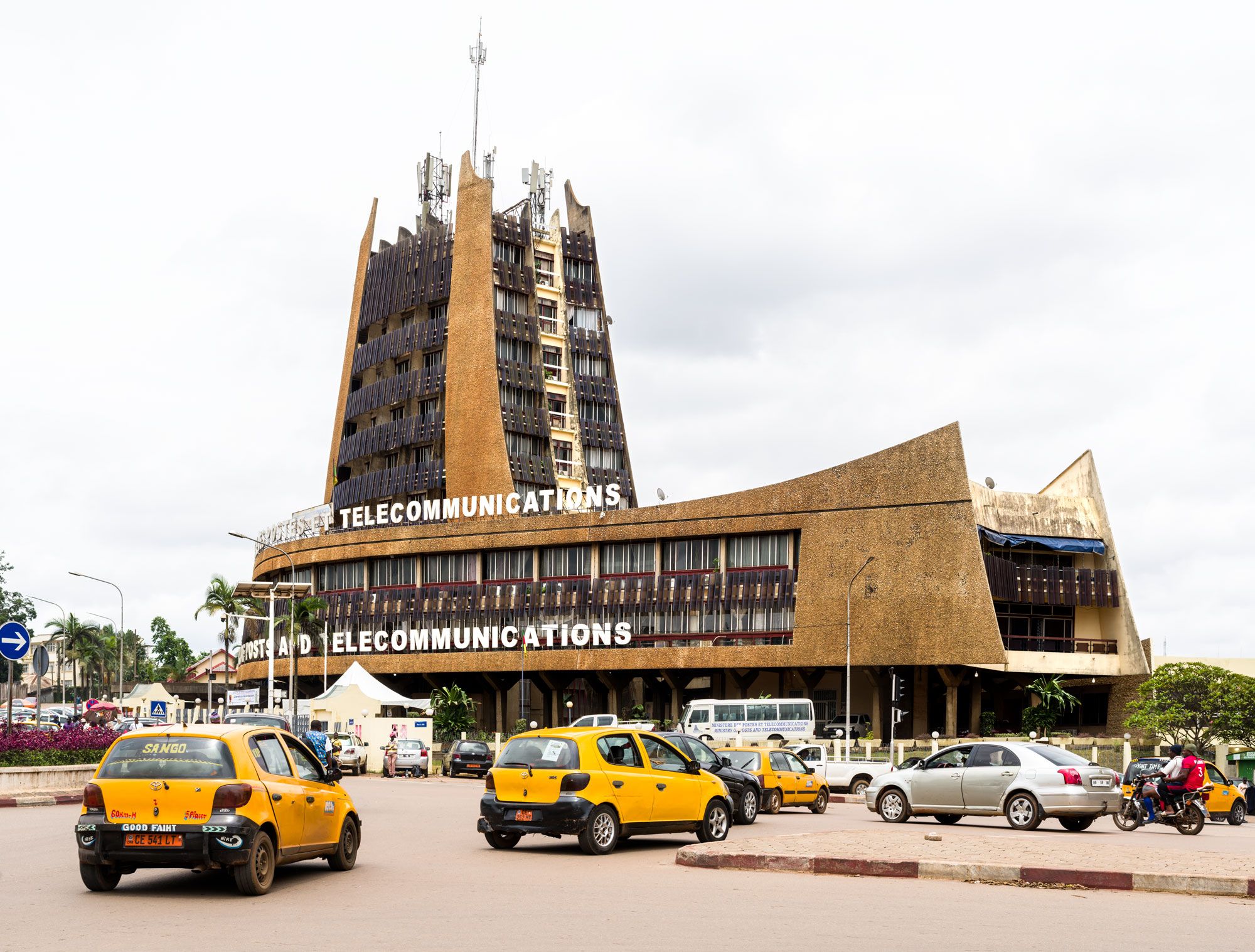 Brutalist telecommunications building with a tall central tower, angular concrete forms, and cars driving through a busy roundabout in front.