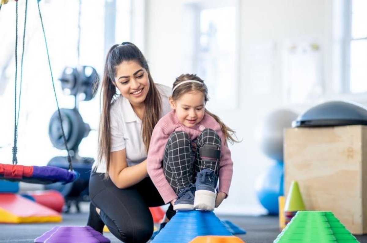 Therapist guiding a smiling girl across balance stepping stones in a therapy gym