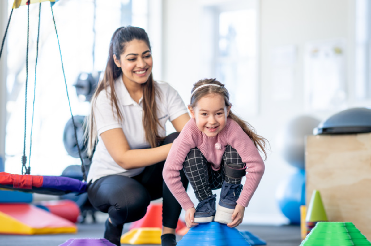Therapist guiding a smiling girl across balance stepping stones in a therapy gym
