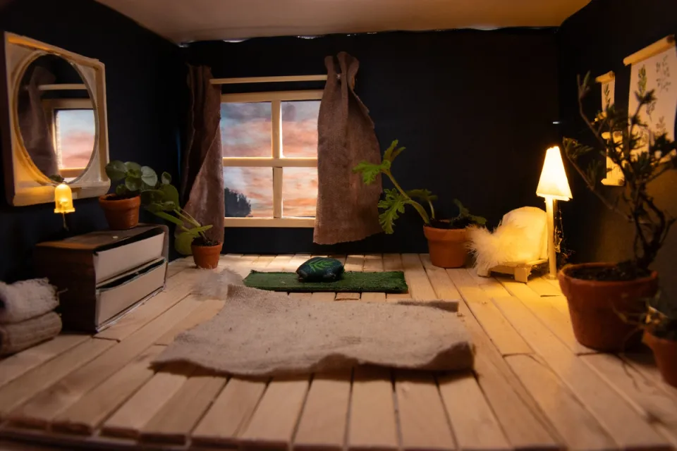 Miniature room inside a shoebox ("kijkdoos"), lit by a warm lamp, with tiny houseplants, a yoga mat, and a wooden floor