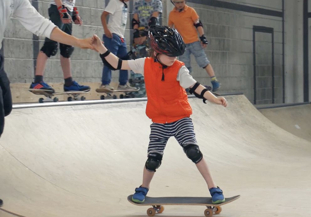 Een kind met helm staat op een board en houdt de hand van de begeleider vast.