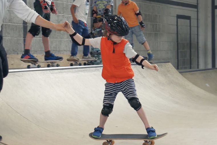 Een kind met helm staat op een board en houdt de hand van de begeleider vast.