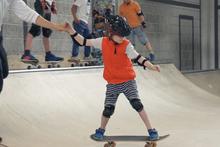 Een kind met helm staat op een board en houdt de hand van de begeleider vast.