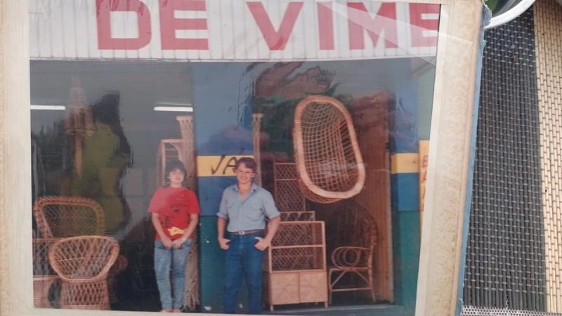 Duas pessoas jovens em frente à fachada de uma loja de móveis de vime, entre poltronas e cadeiras.