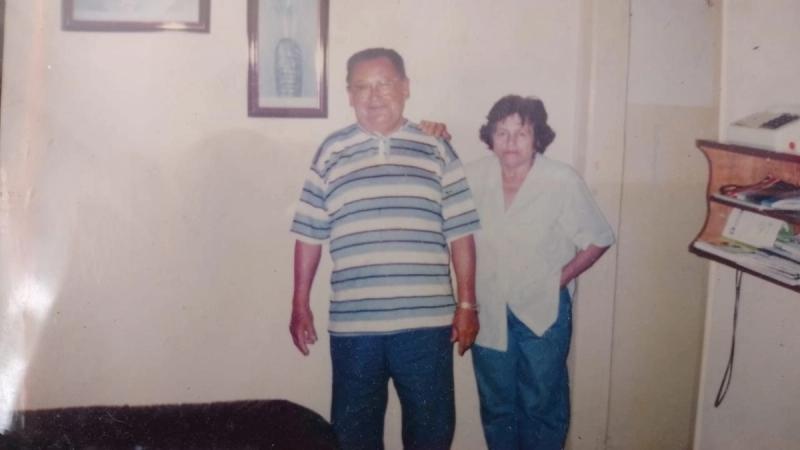 Um casal posa lado a lado em frente a uma casa simples, nos fundos de uma loja.