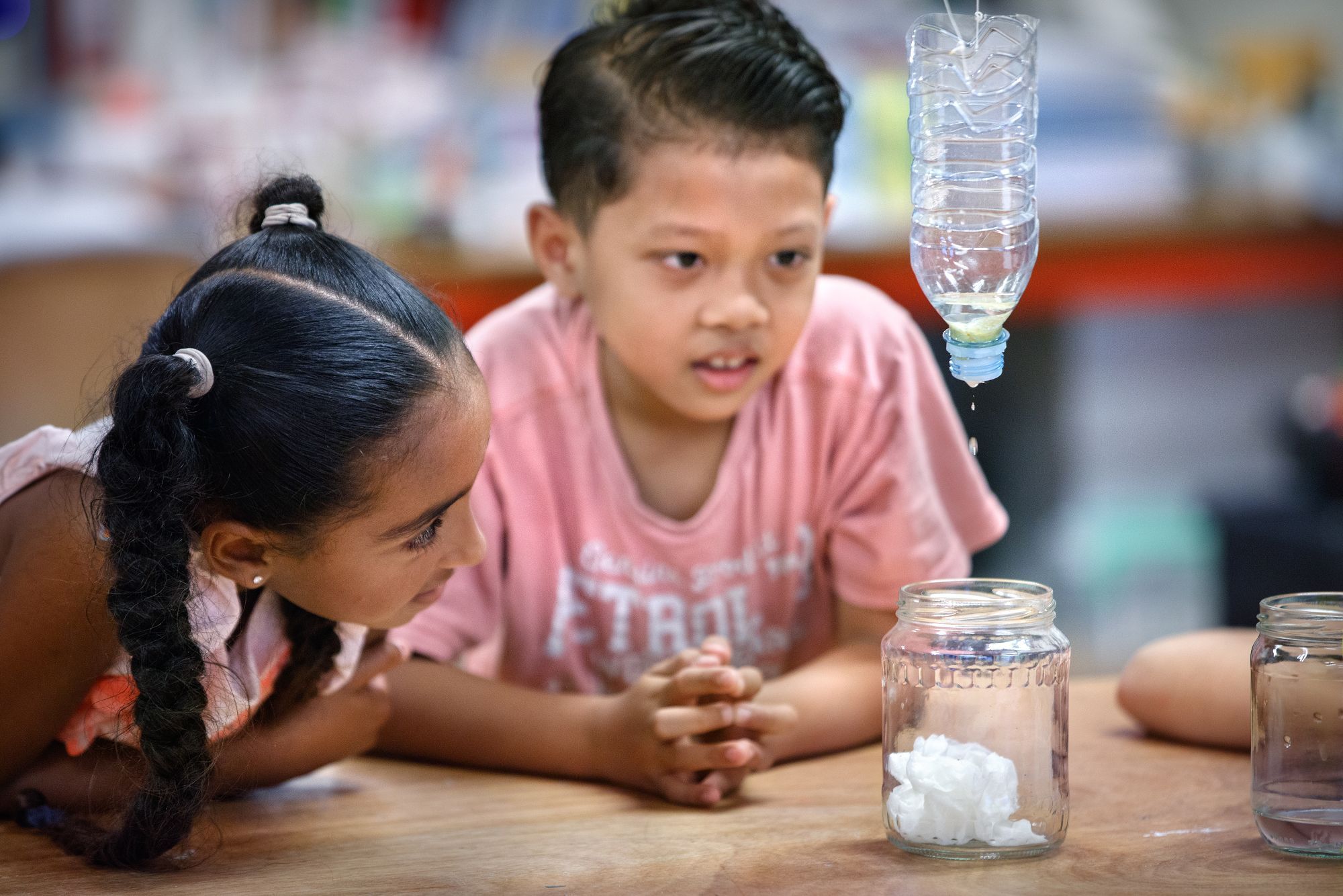 Een jongen en een meisje kijken toe hoe er water uit een fles in een potje druppelt.