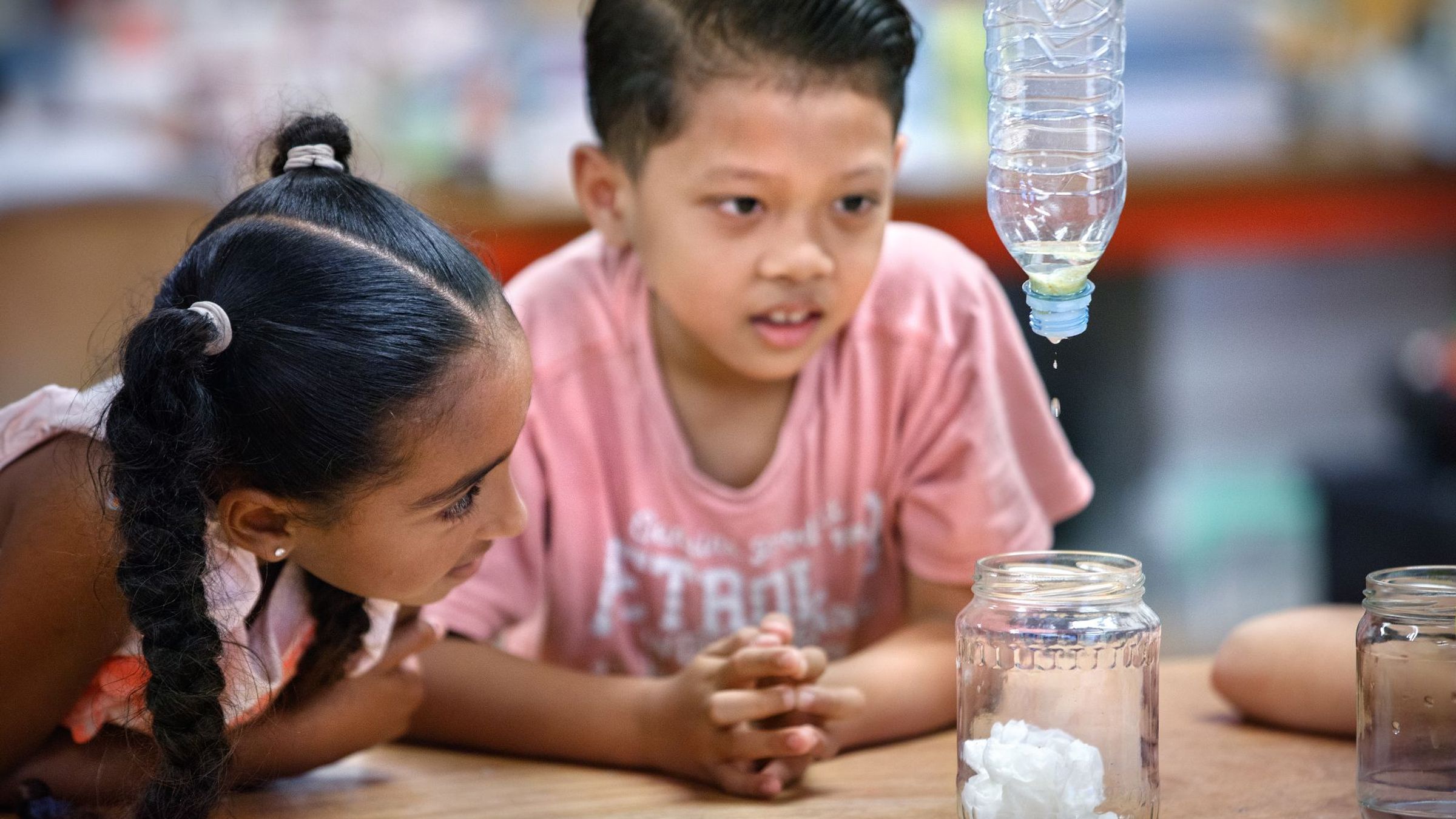 Een jongen en een meisje kijken toe hoe er water uit een fles in een potje druppelt.