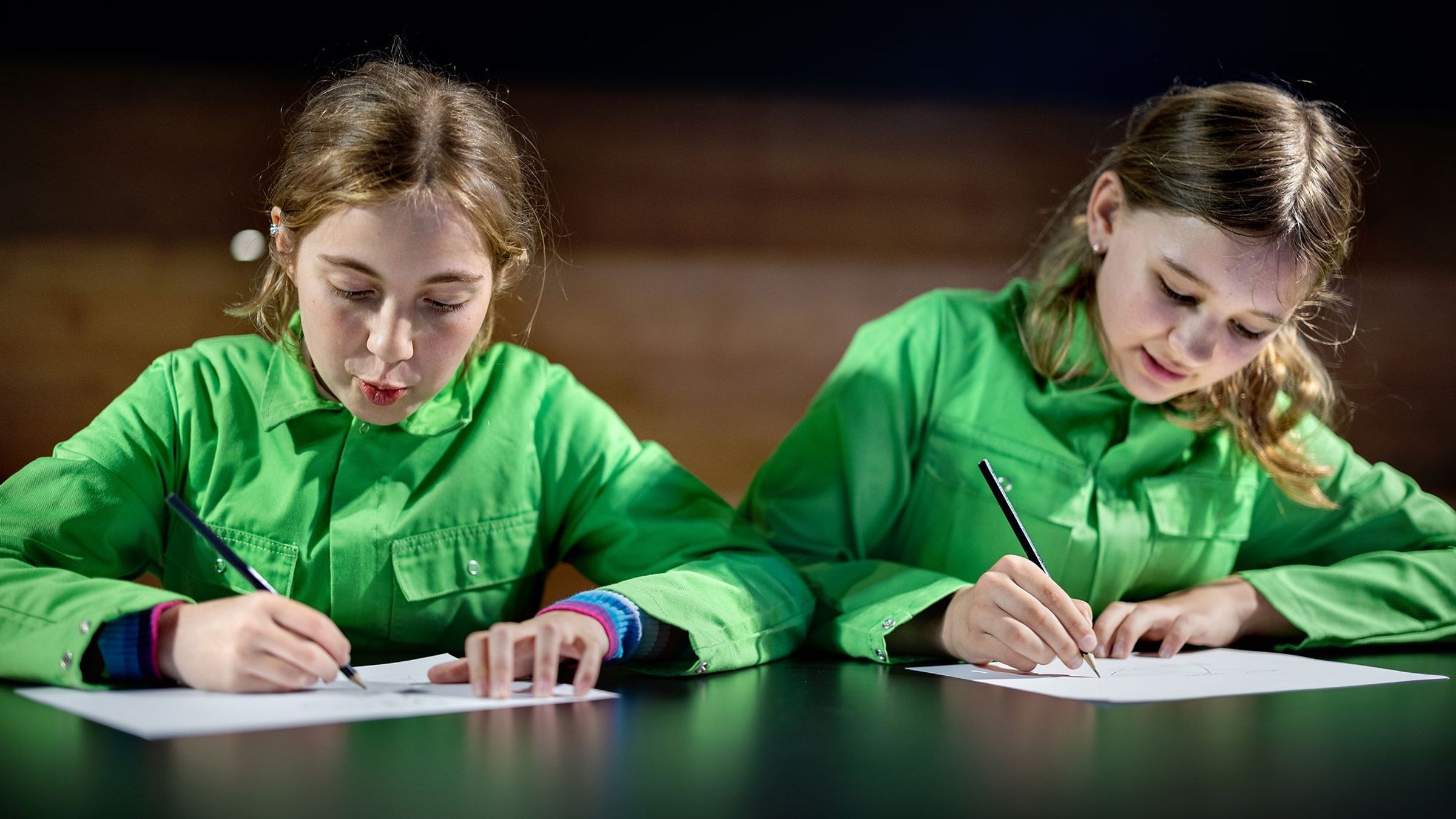 Twee meisjes in groene pakken tekenen op een blaadje.