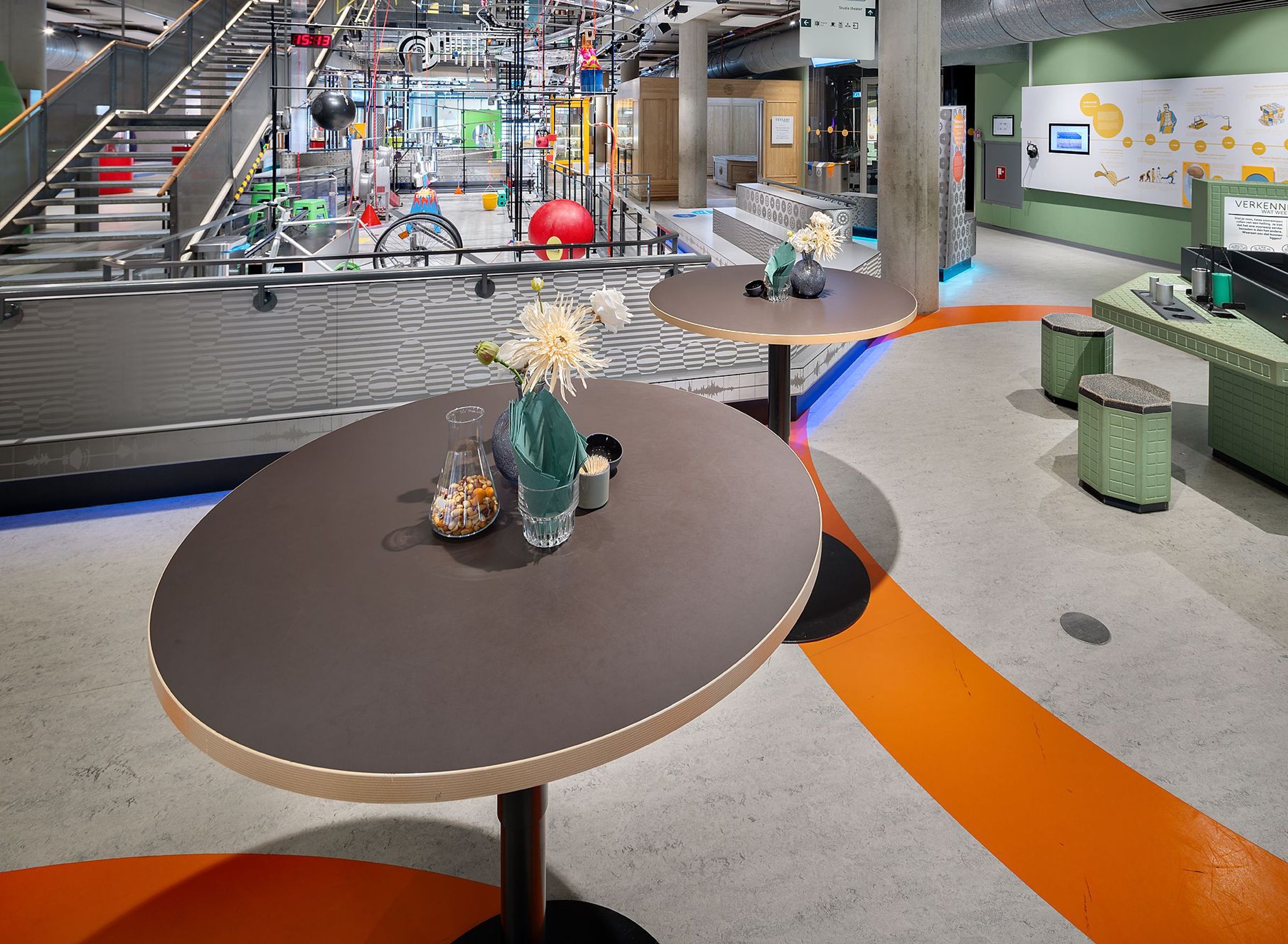 Colourful exhibitions, with orange circles on the floor. Two tall round tables set with flowers and snacks.