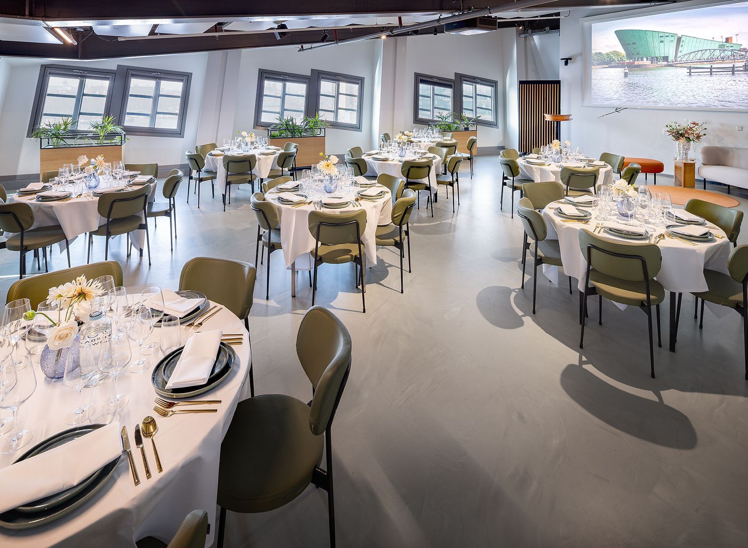 Een zaal met grijze vloer, witte ronde tafels en groene stoelen.