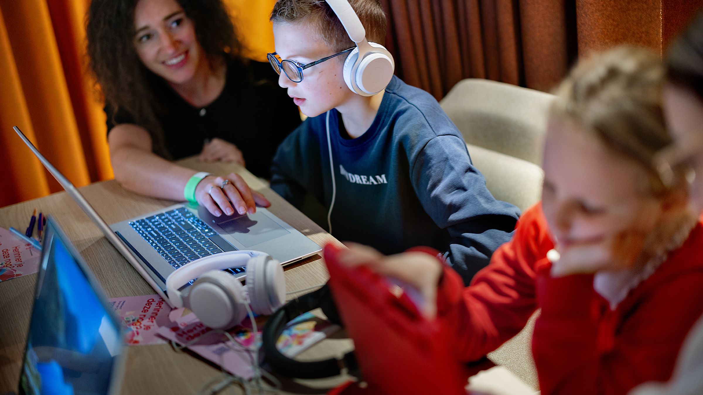 een vrouw wijst iets aan op een laptop, kind met koptelefoon kijkt