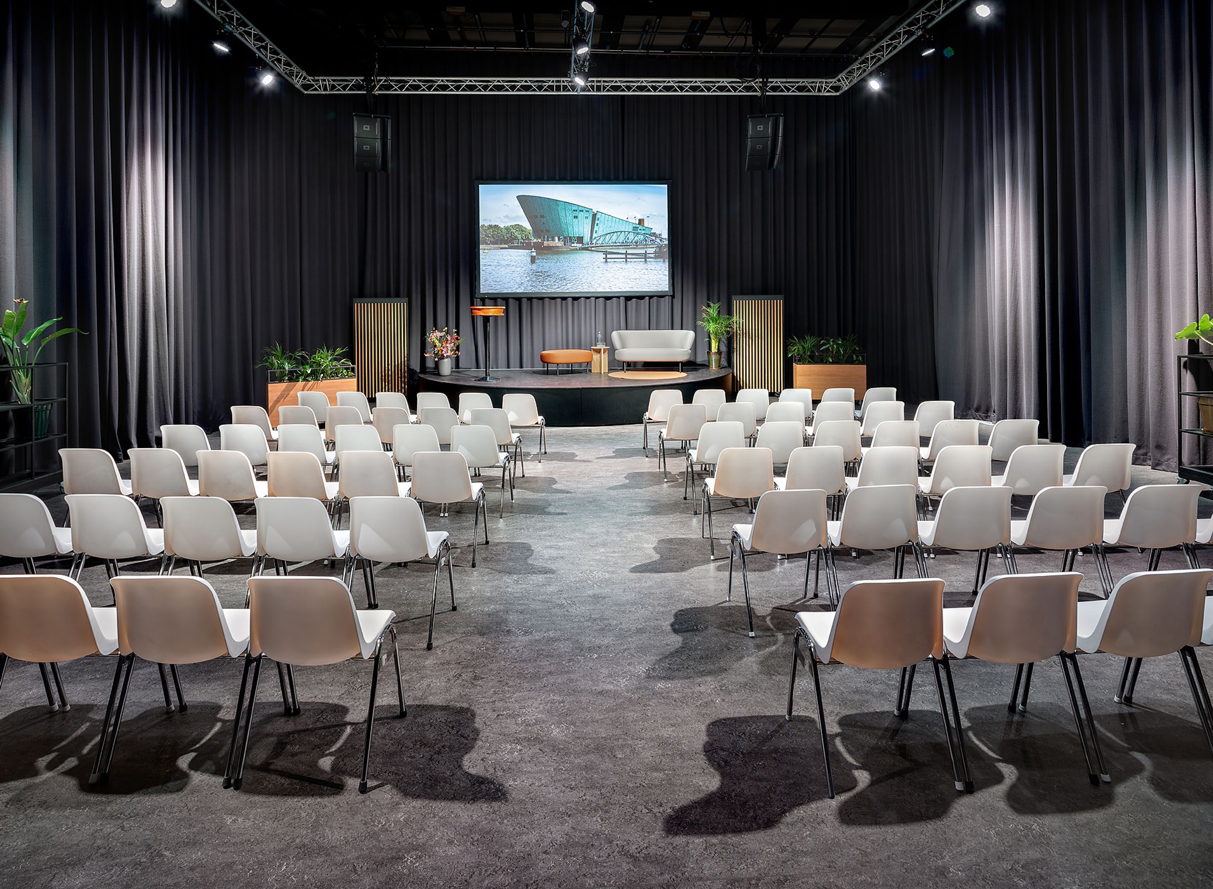 Een grote grijze zaal met witte stoelen.