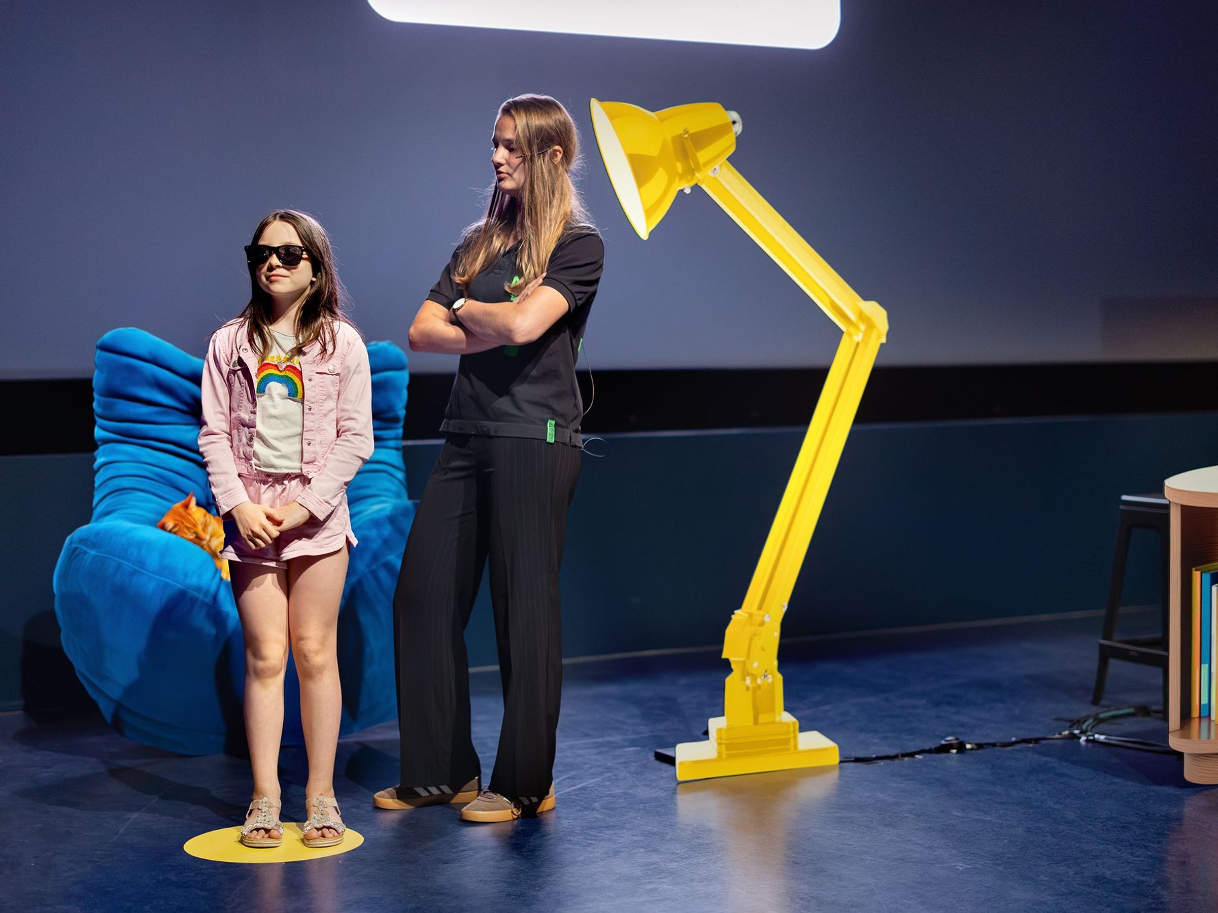Een meisje met een zonnebril en een vrouw met haar armen over elkaar staan naast elkaar, met een grote gele lamp naast zich