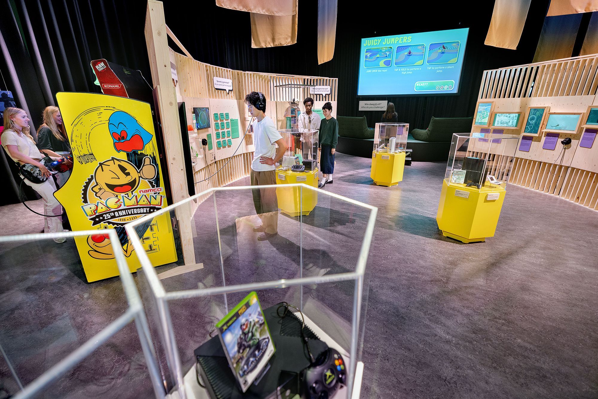 People are looking at different objects in an exhibition, with wooden panels and game consoles in glass display cabinets.