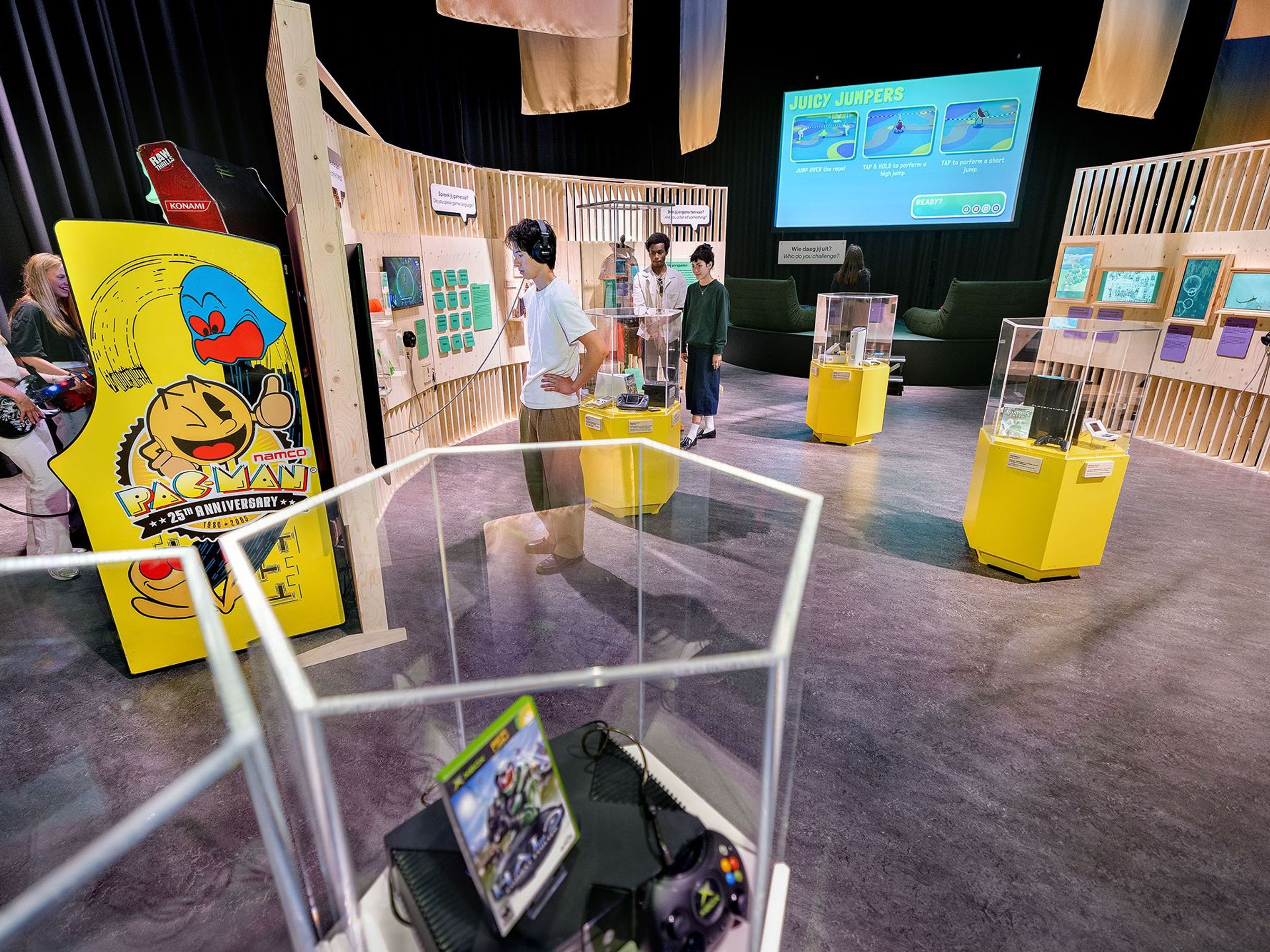 Mensen staan in een tentoonstellingsruimte met houten panelen en game consoles in vitrines