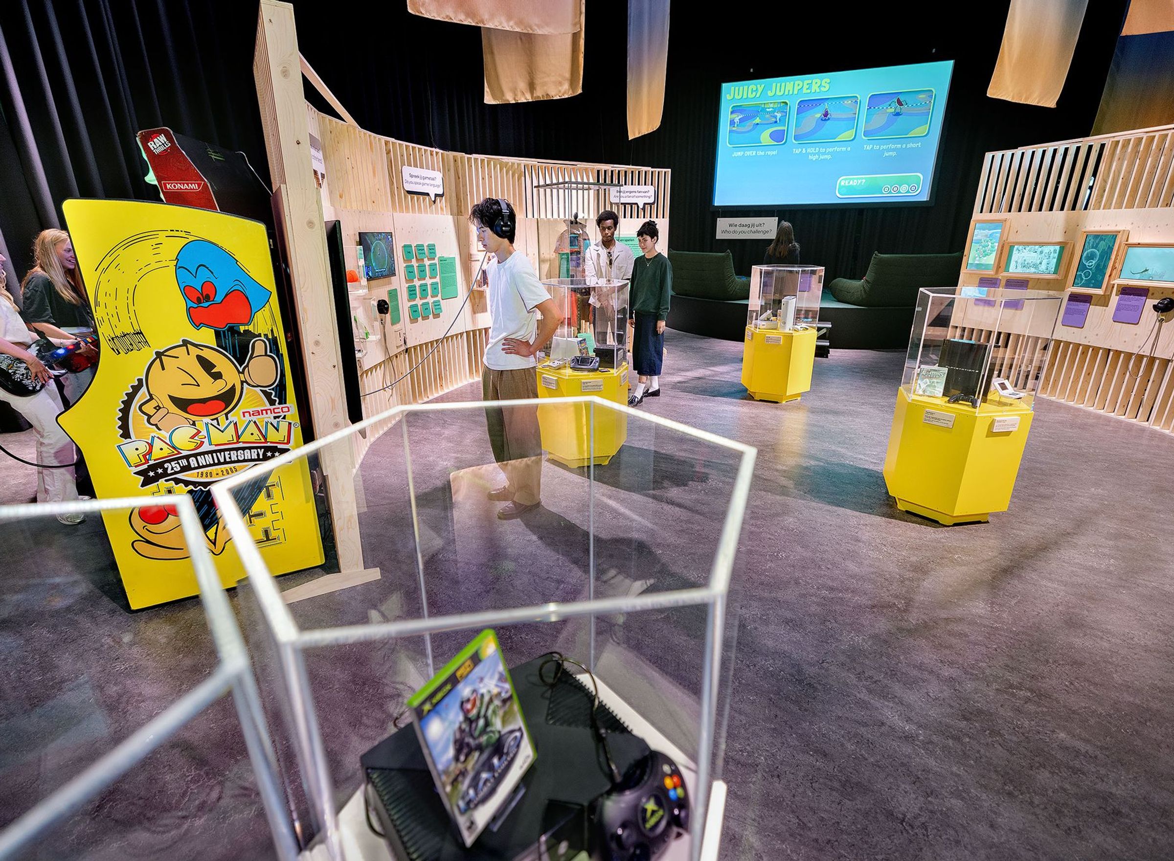 People are looking at different objects in an exhibition, with wooden panels and game consoles in glass display cabinets.