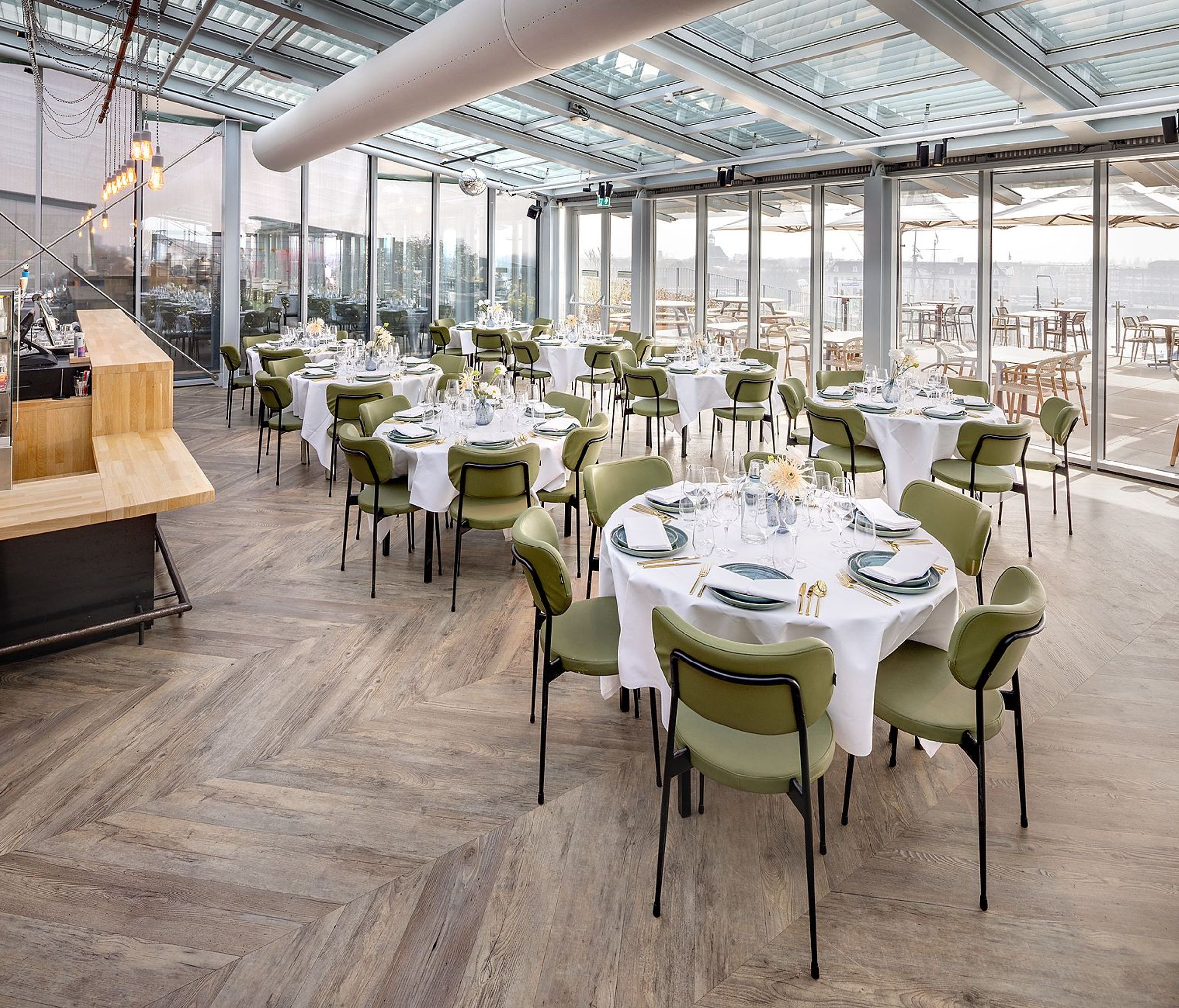 Een ruimte met een houten vloer, veel glas en ronde gedekte tafels met groene stoelen.