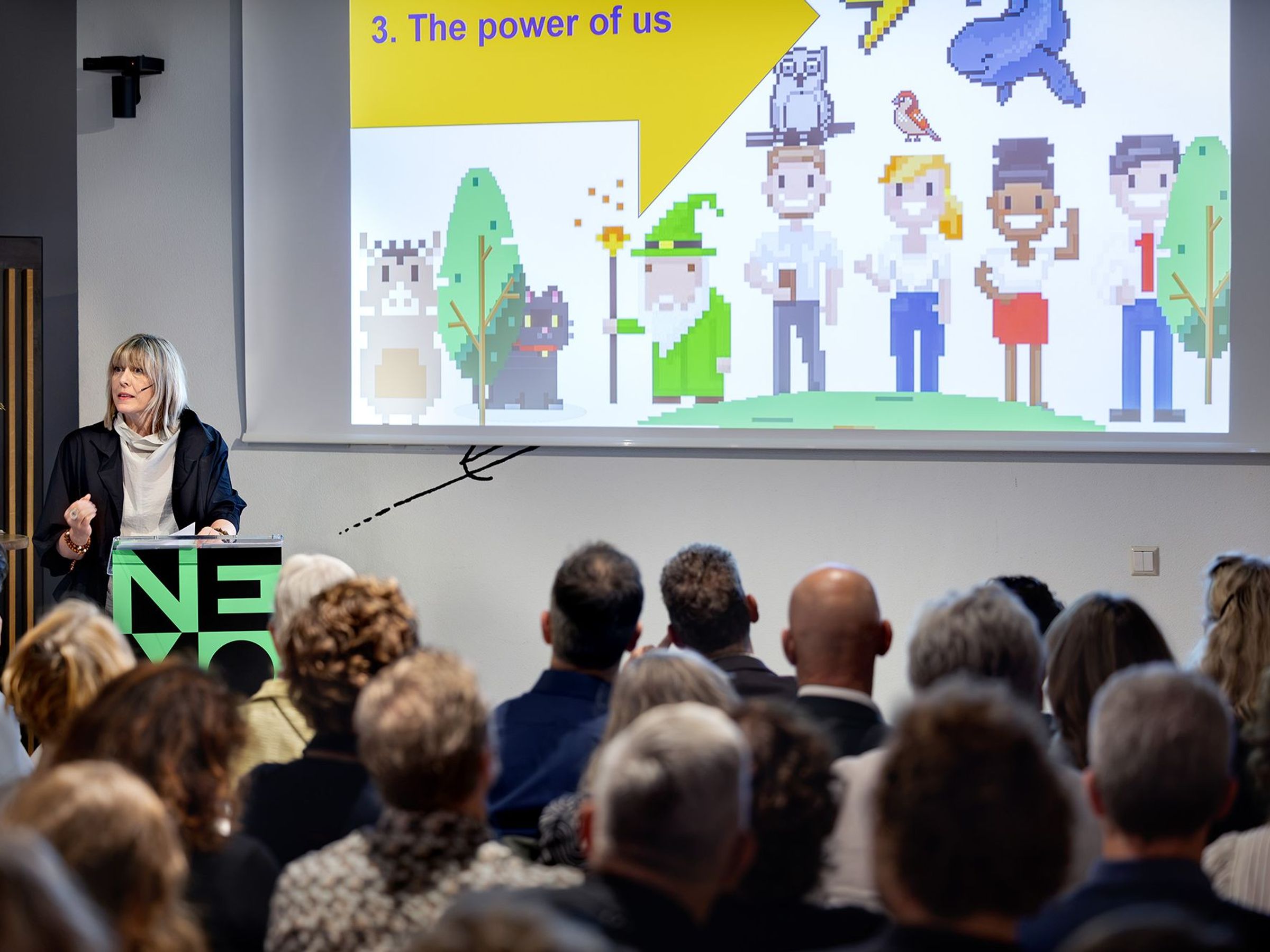 Een vrouw staat achter een spreekgestoelte en spreekt een groep mensen toe