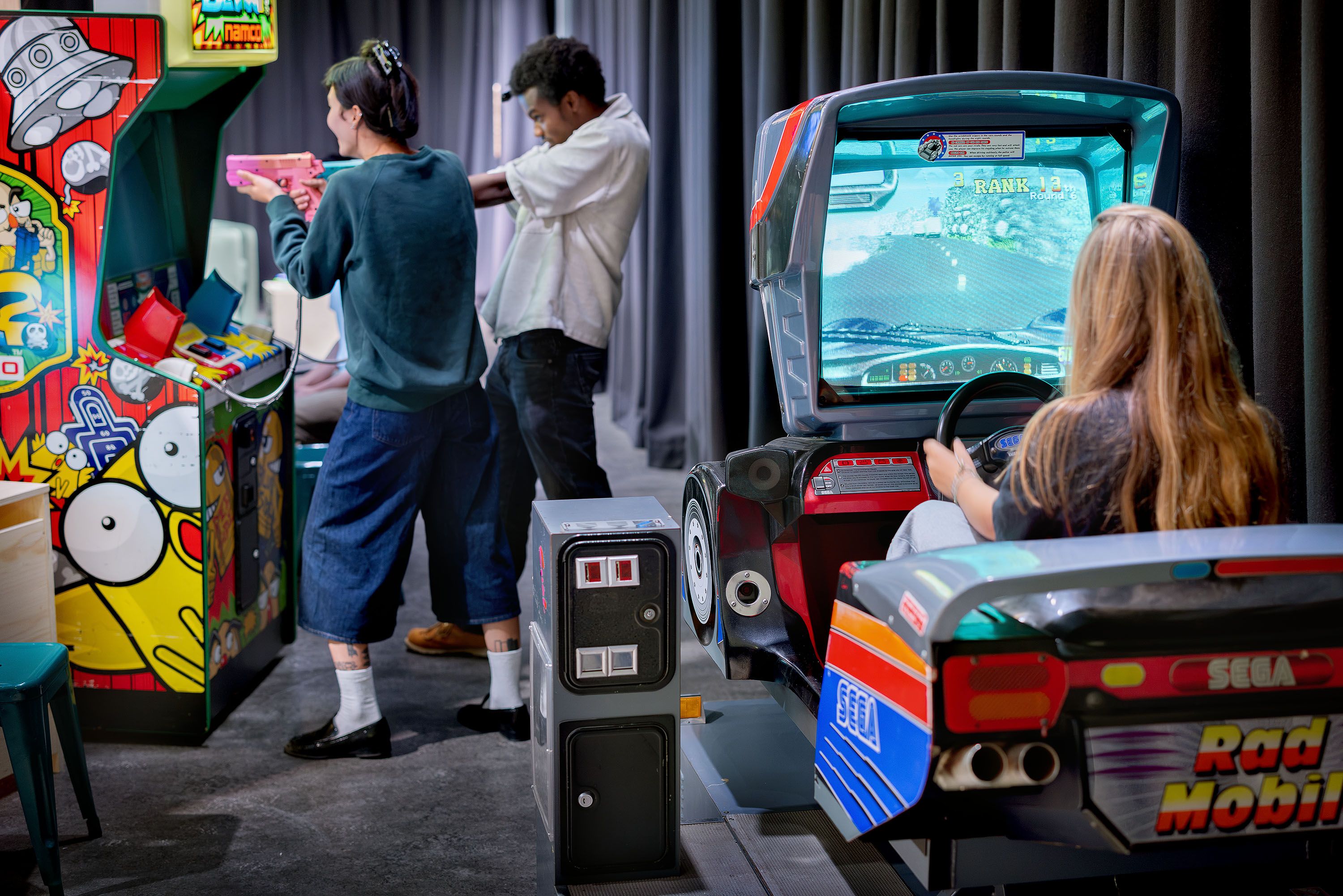 A girl is sitting in a car in a video game, with a boy and a girl in the background shooting at a screen with toy guns.