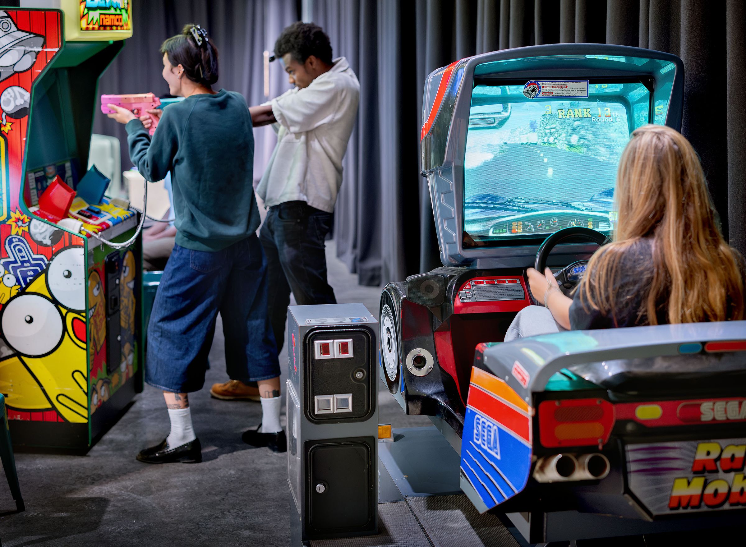 A girl is sitting in a car in a video game, with a boy and a girl in the background shooting at a screen with toy guns.