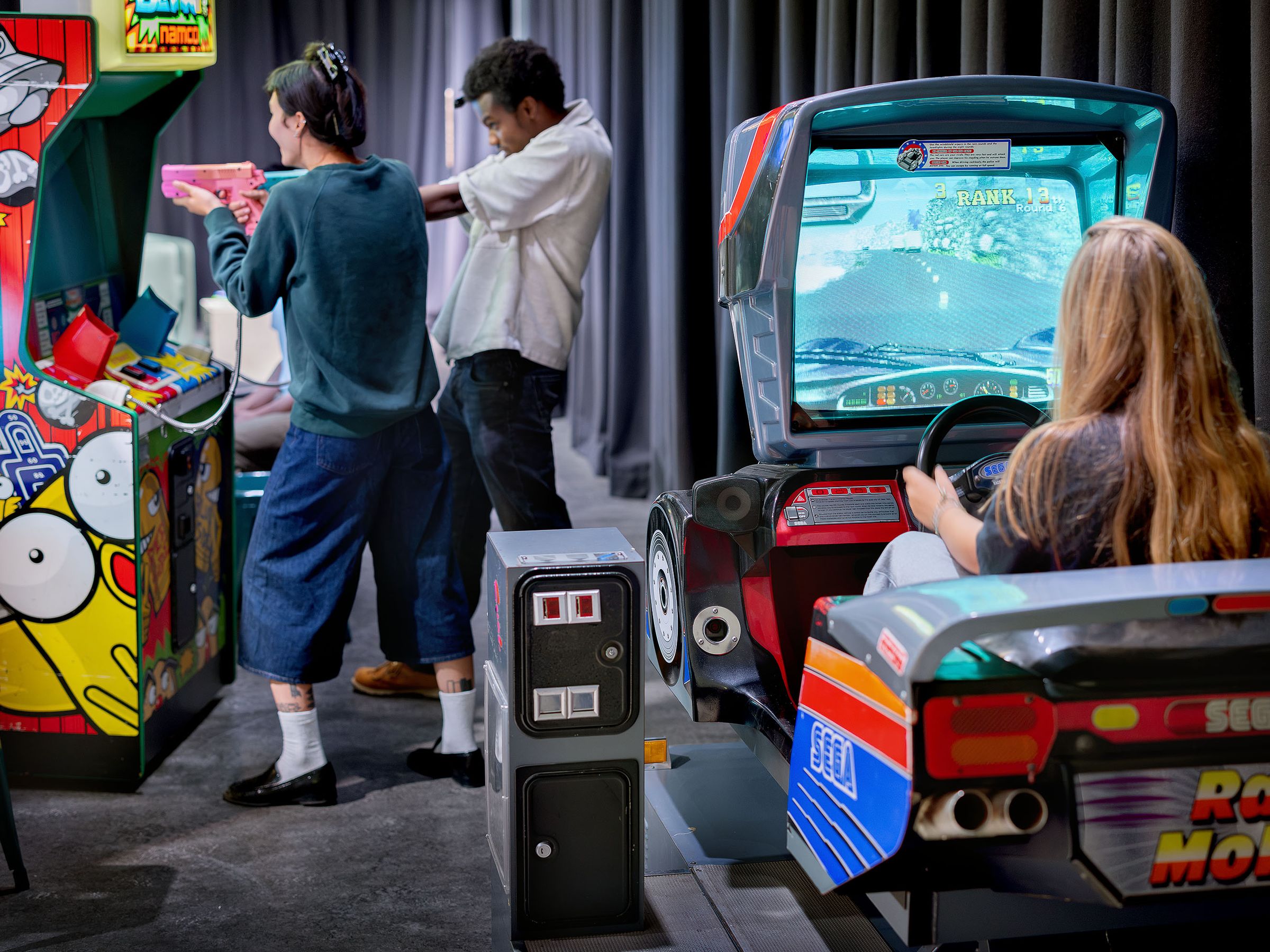 Een jongen en een meisje staan bij een oude arcadegame met speelgoedwapens, een meisje zit in arcadegame in de vorm van een auto