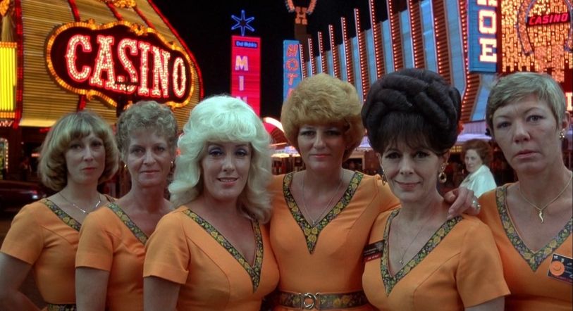Zes vrouwen poseren in oranje jurken in het casino in de jaren 80.