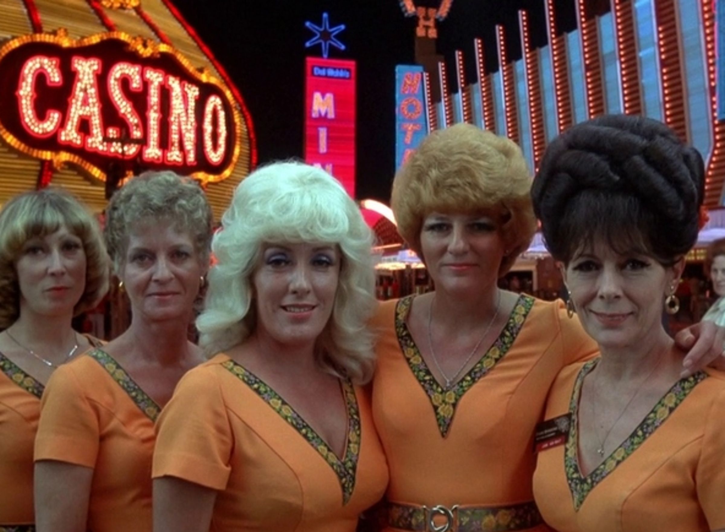 Zes vrouwen poseren in oranje jurken in het casino in de jaren 80.
